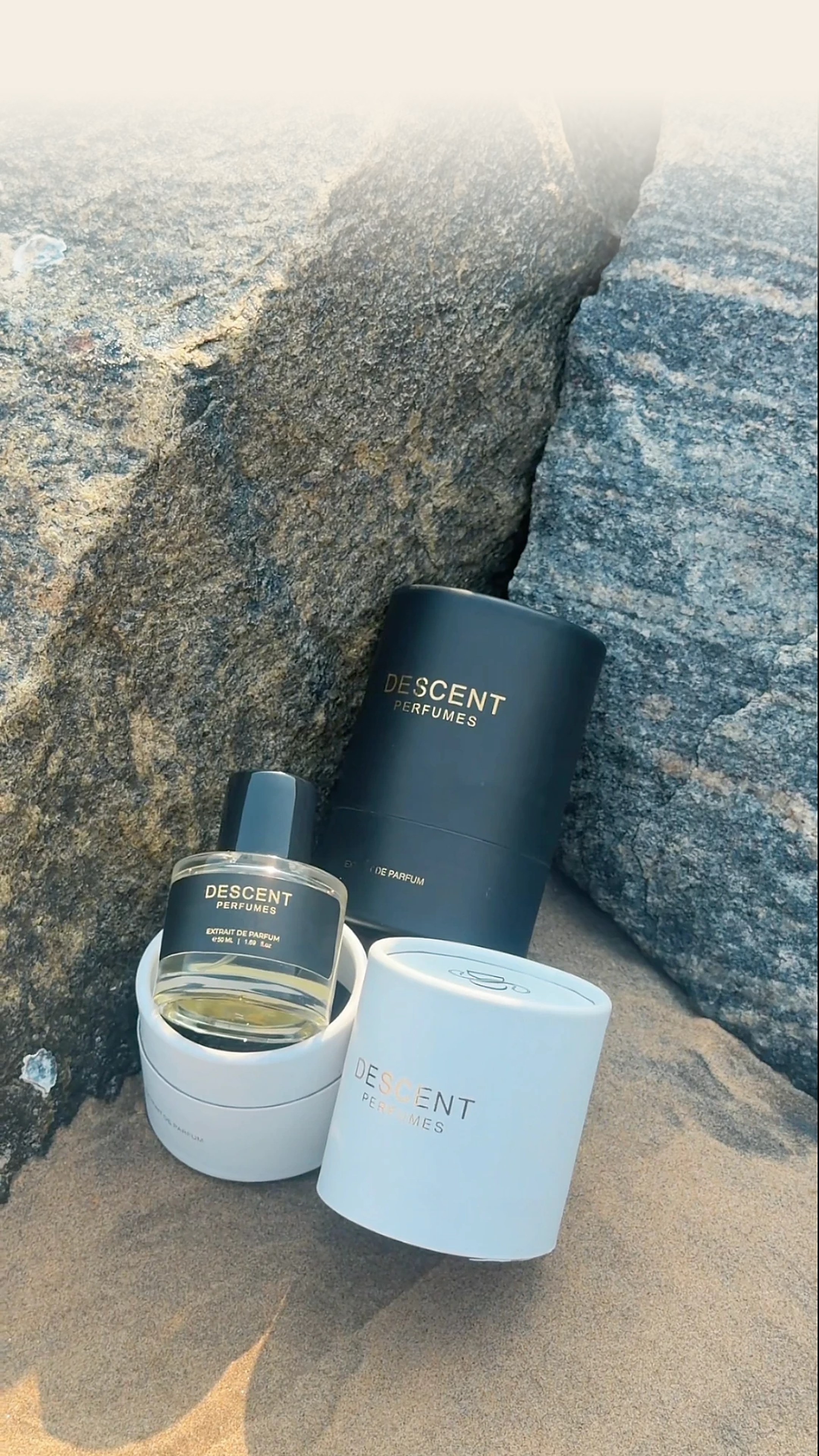 The Descent extrait de parfum resting in its white keepsake tube beside the black and white gift tubes, on golden sand between coastal rocks